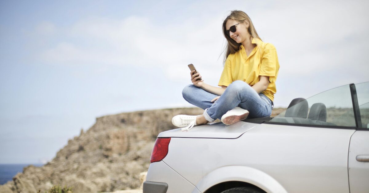 Young Caucasian woman sitting on the hood if a silver convertible, wearing sunglasses, on her phone