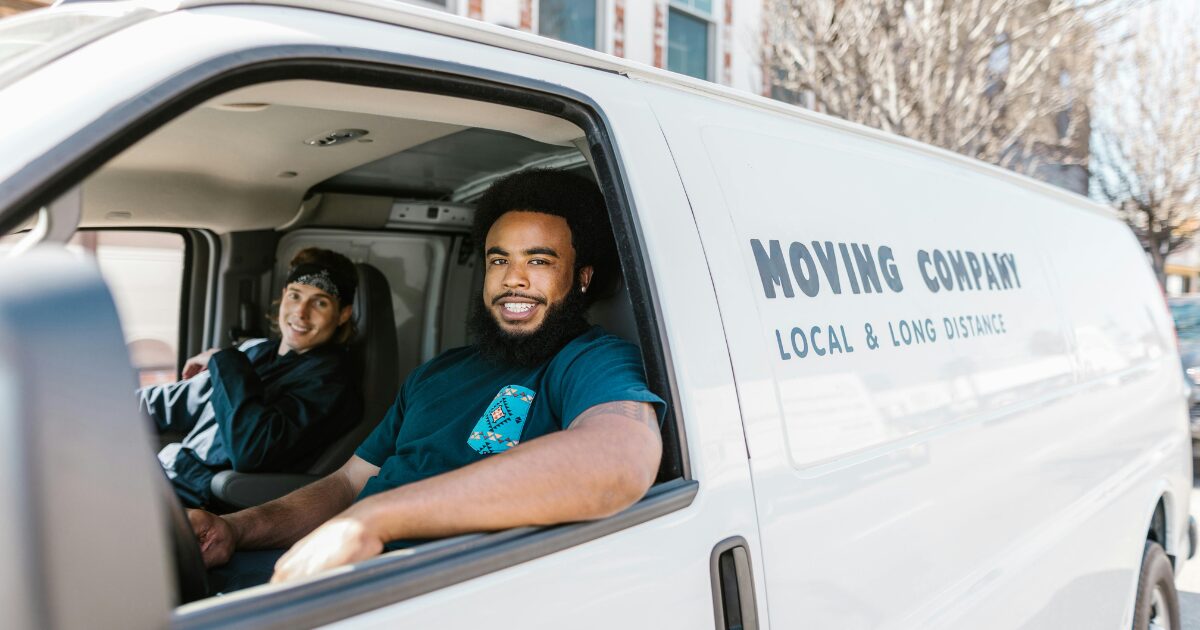 A Black man driving a white company van, with a Caucasian man in the passenger seat.