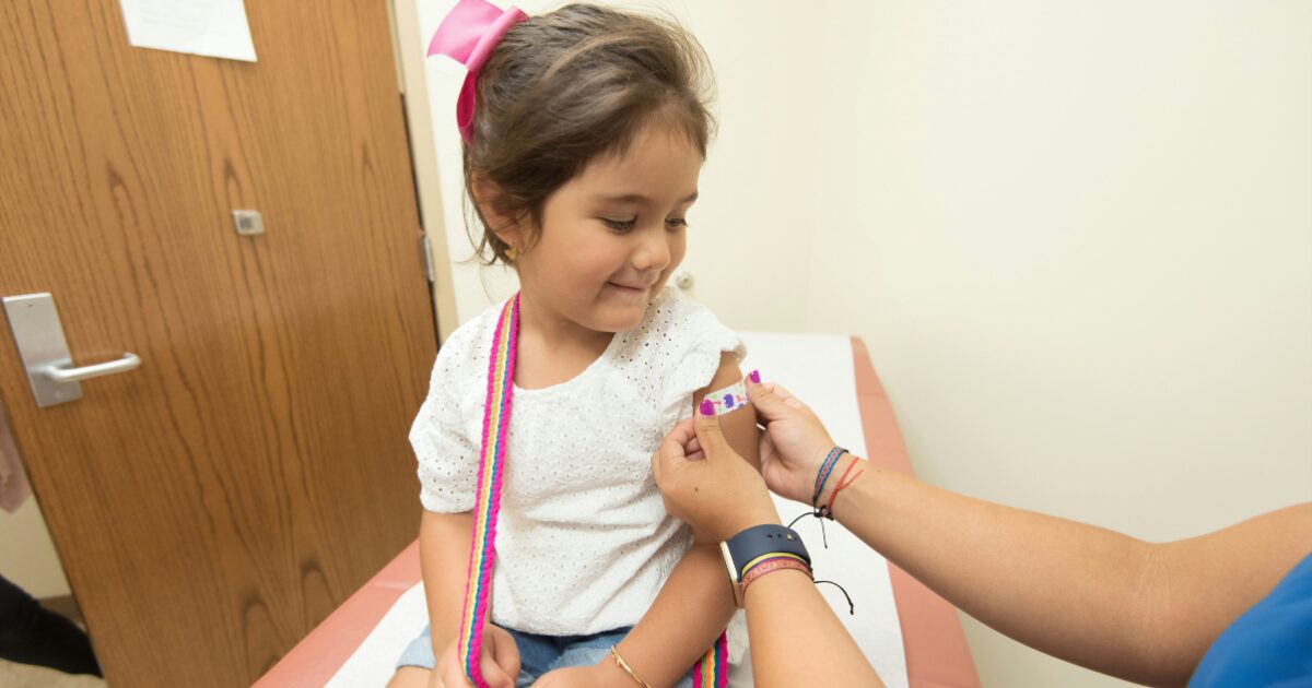 A young Hispanic girl getting a band-aid put onto her arm after a vaccine.