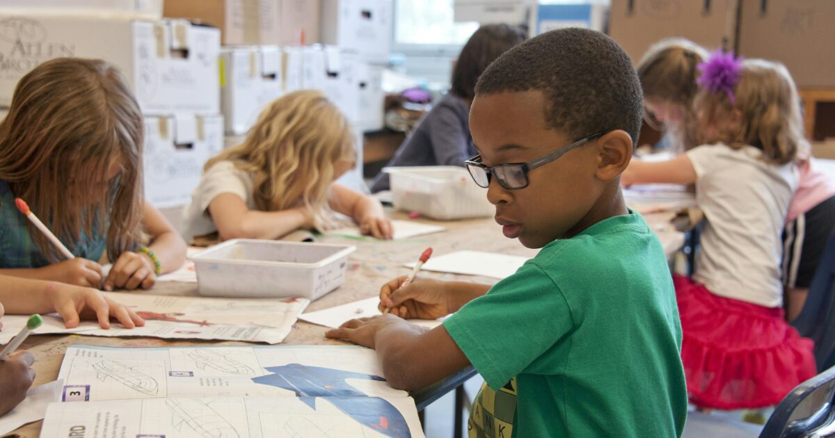 Young children writing in a classroom, with a little Black boy in a green shirt closest to the camera.