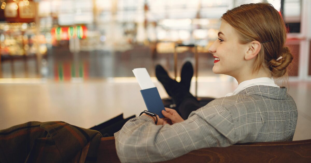 Young woman siting in an airport wearing a blazer, with her passport in her hand, smiling