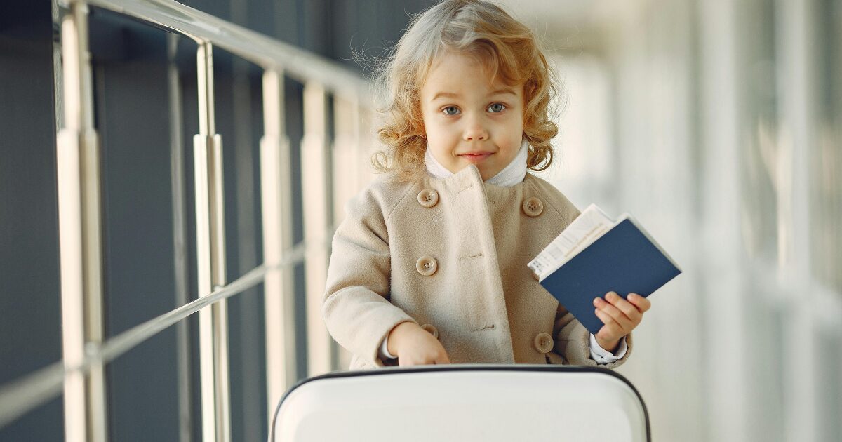 Young girl with blonde curls and a beige coat, holding her passport.