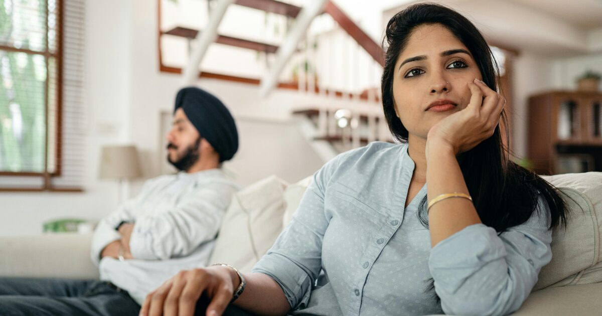A South Indian couple (male and female) sitting on a couch in a brightly lit home. Both are looking in different directions, looking upset
