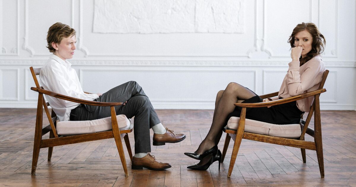 A young couple Caucasian couple (male and female) sitting across from each other in wooden chairs. They look visibly upset, the woman is looking to the side