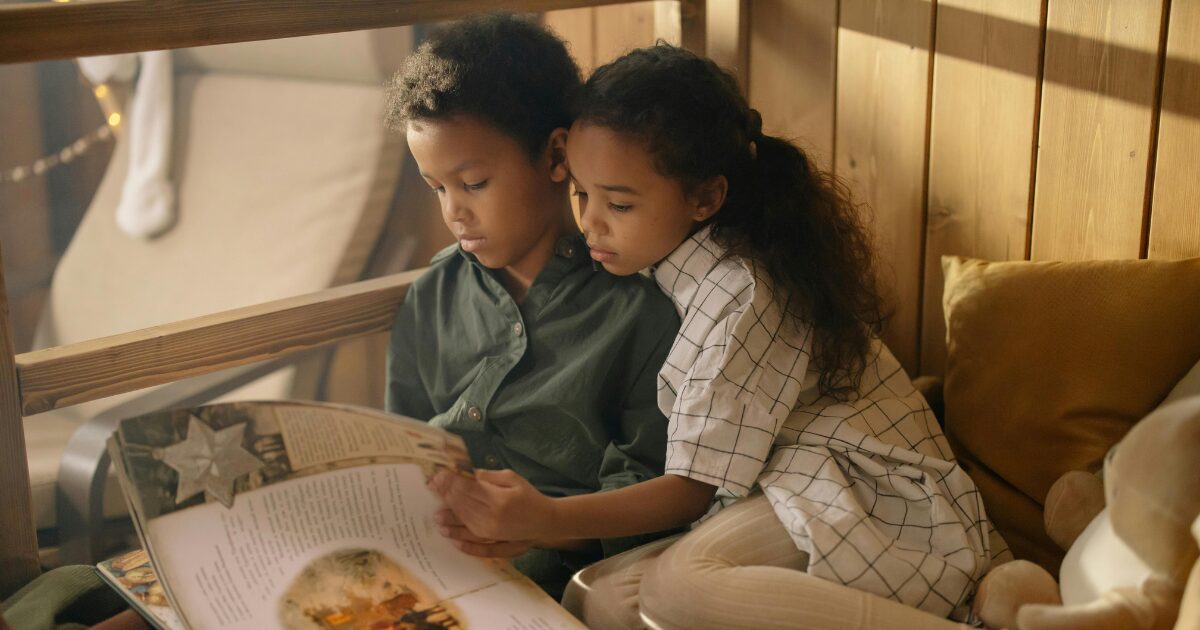 Two young Black children sitting in a wooden sunroom reading a book together.