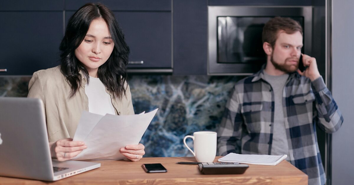 An East Asian woman reading over paperwork at a wood kitchen table and Caucasian man on the phone in the background.