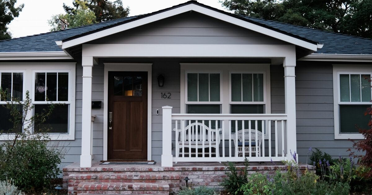 A single family home with light gray siding, a dark gray roof, a white porch, and a wooden door.