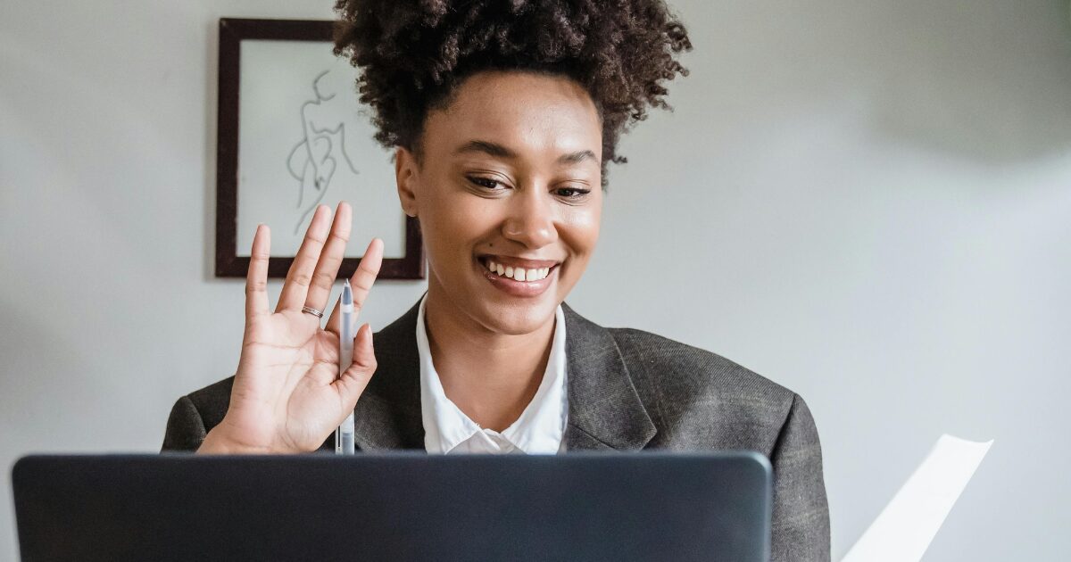 A young Black woman on a video call smiling while holding paperwork and waving to the camera.