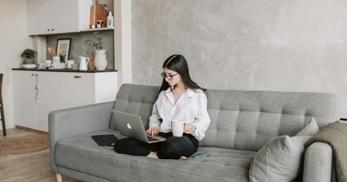 A young Caucasian woman sitting on a grey couch, working on her laptop