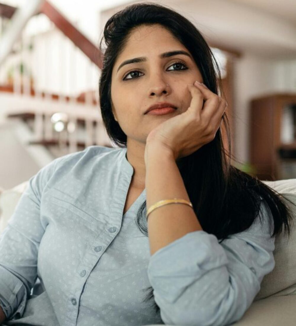 A South Indian female sitting on a couch in a brightly lit home. She is looking concerned, as if she is in deep thought.