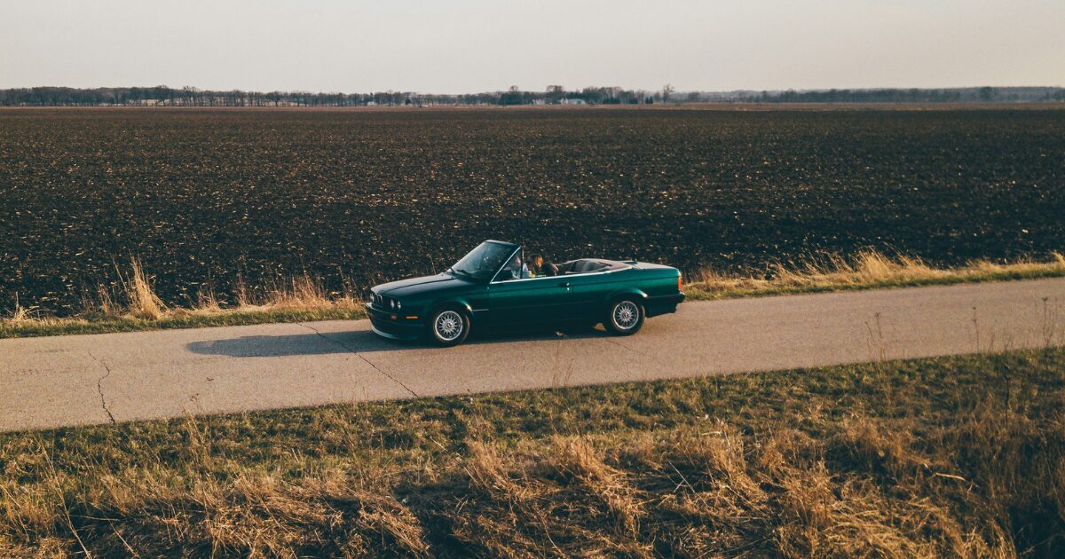 A forest green convertible driving down an open country road.