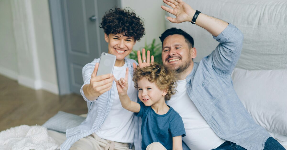 A Caucasian mom and dad sitting on their bed with their toddler, waving to someone on a mobile video call.