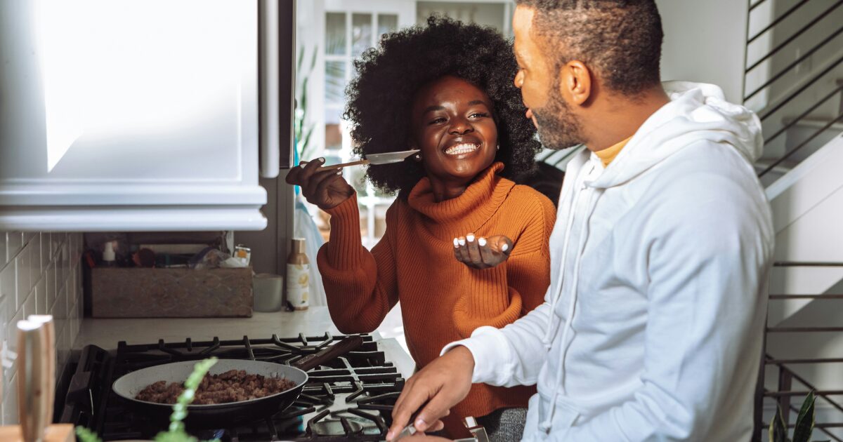 A middle-aged Black couple cooking together, with the woman giving him food to try on a serving spoon.
