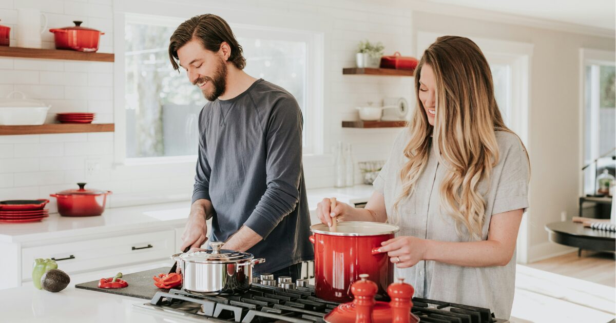 A Caucasian couple (male and female) standing in a bright white kitchen, cooking together.