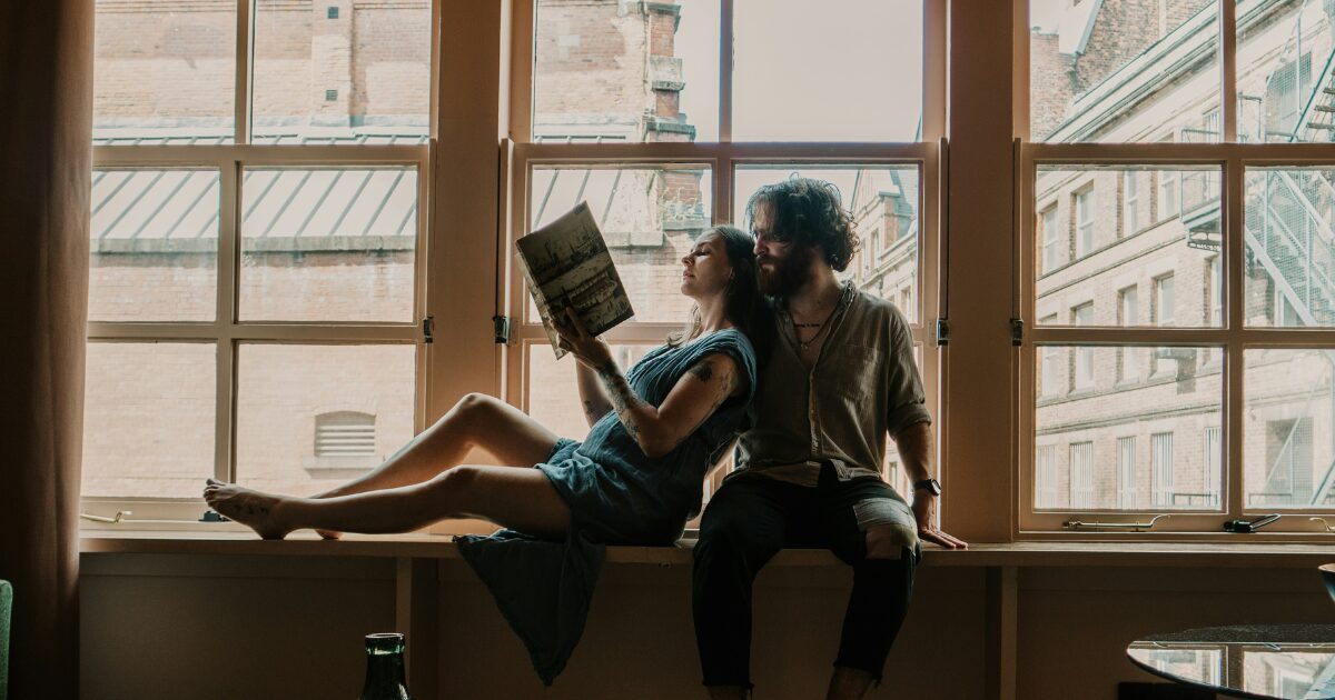 a middle-aged Caucasian couple (a man and woman) sitting on a windowsill, reading a book together.