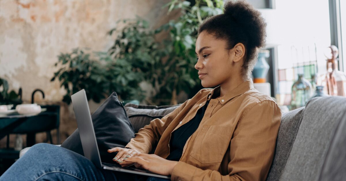 A young Black woman sitting on a couch, working on her laptop.