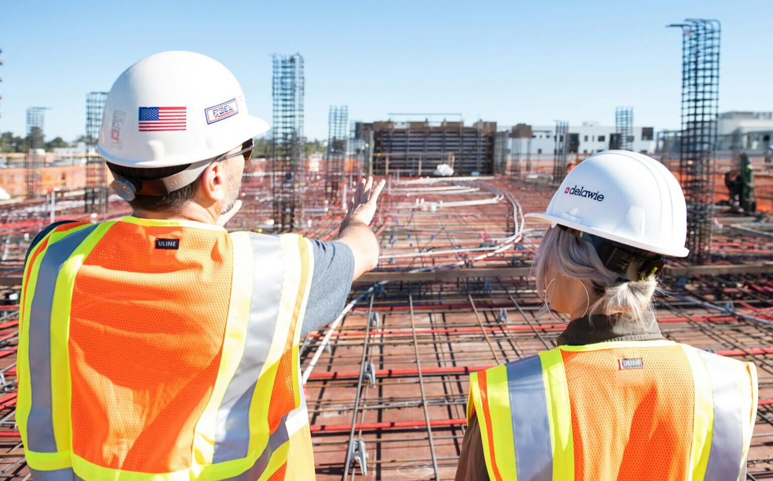 A male and female construction worker with their backs to the camera, looking over a job site. They are wearing high-visibility orange vests and white hard hats.