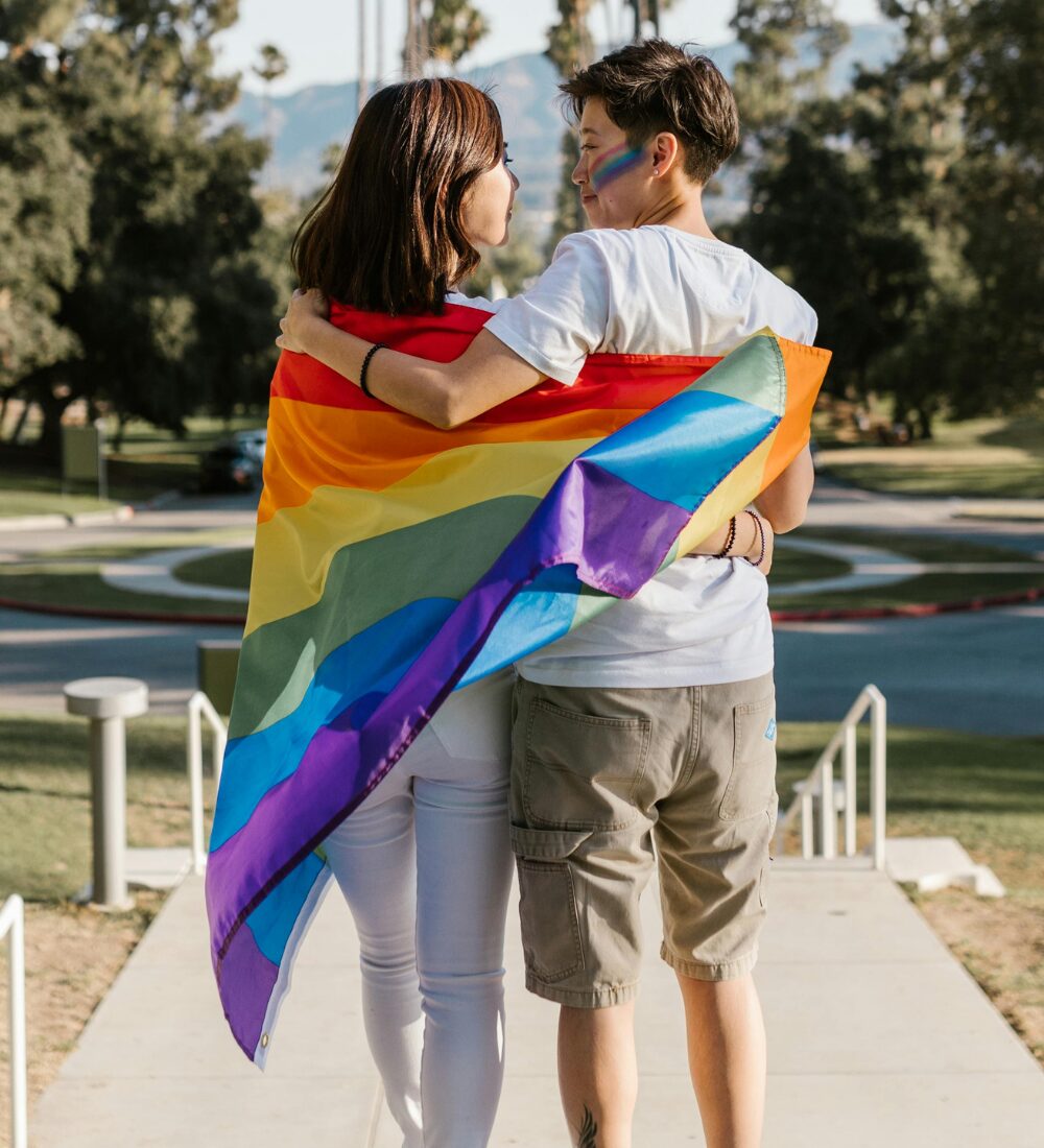 Two East Asian women with their arms around each other, wrapped up in a rainbow pride flag.