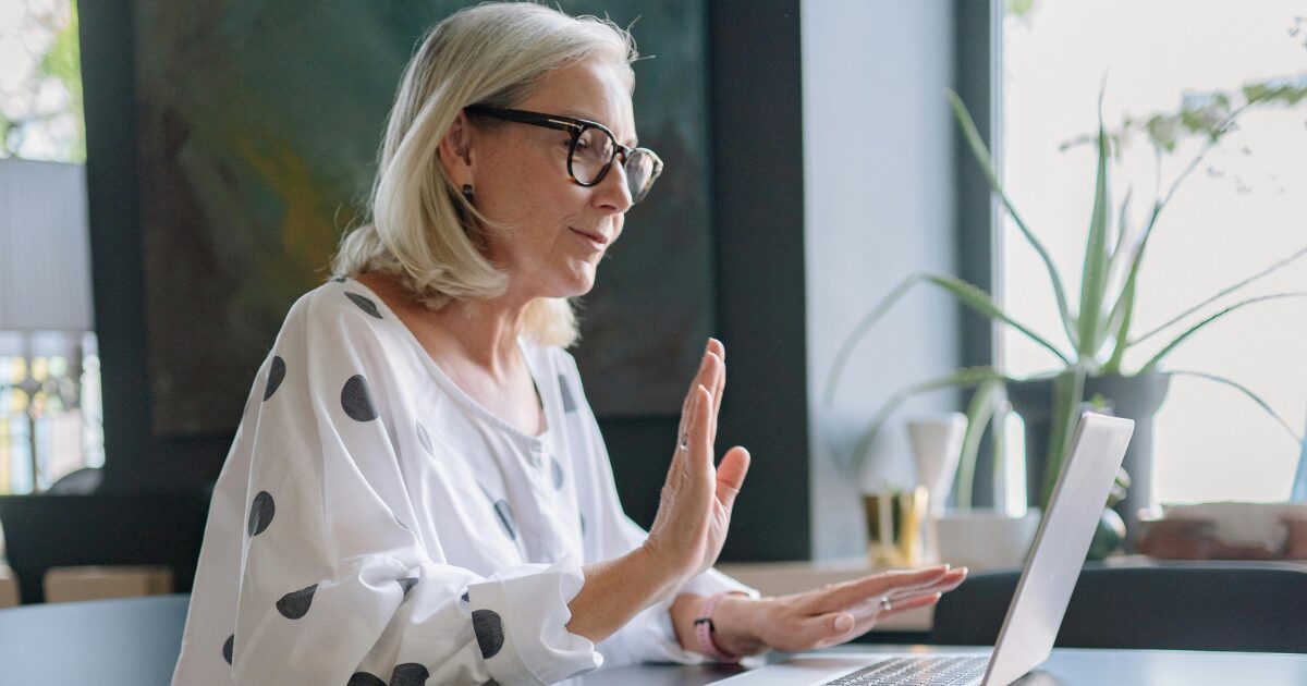 A Caucasian senior woman on a video call on her laptop with her hand up.