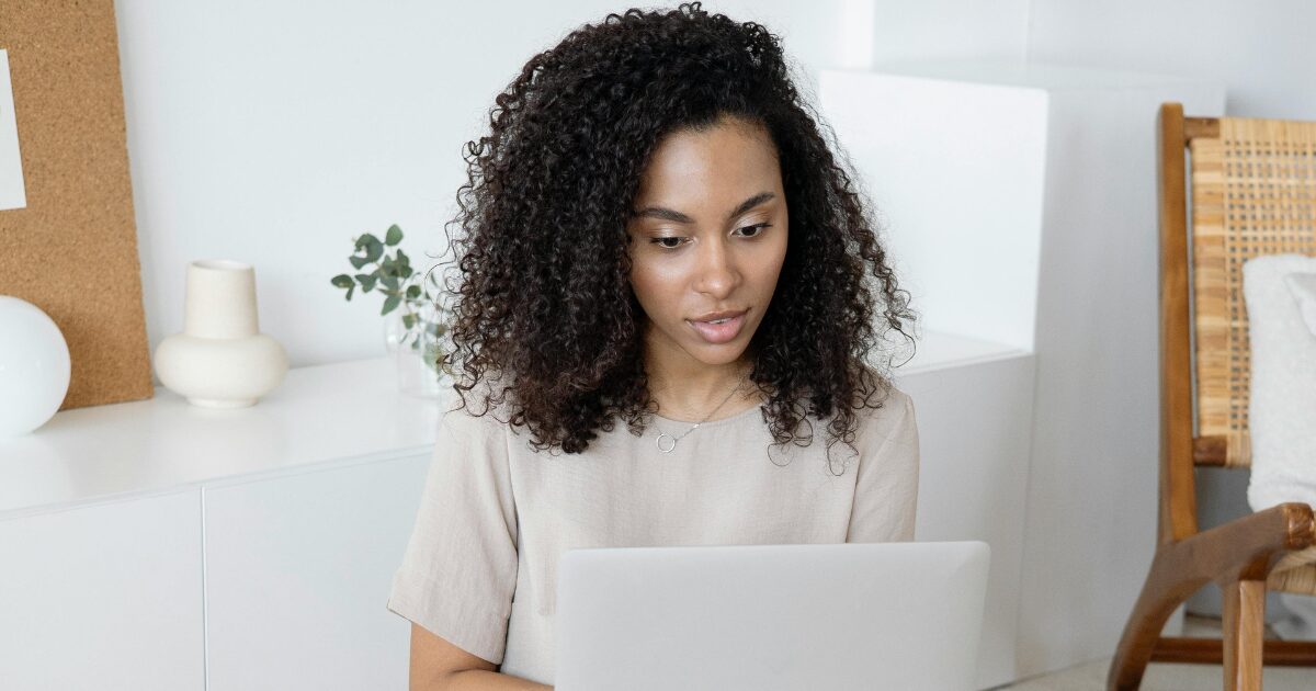 Young Black woman in a beige shirt on her laptop in a living room.