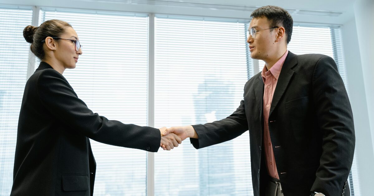 A young East Asian woman shaking hands with an East Asian man. Both are wearing a black blazer.