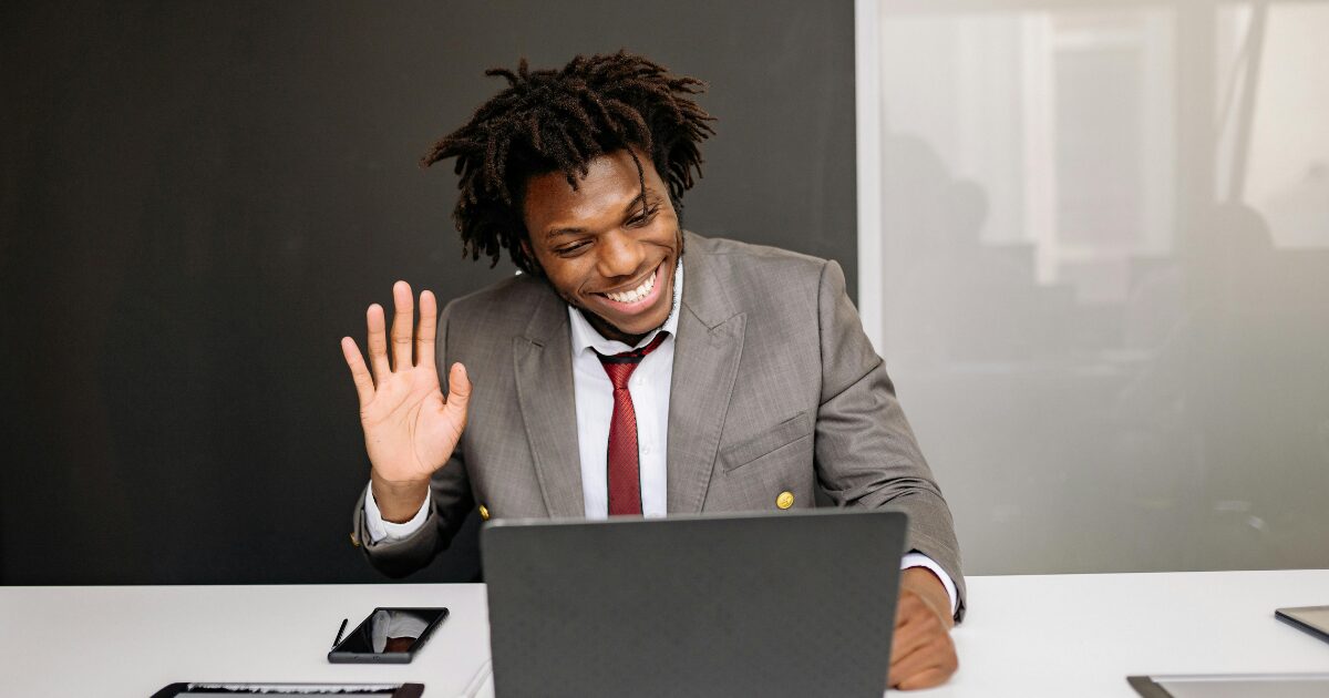 Black adult man in a grey suit, sitting at a table waving to his open laptop.