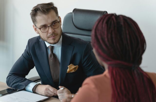 A Caucasian man in a suit talking to a Black woman, her back is facing us