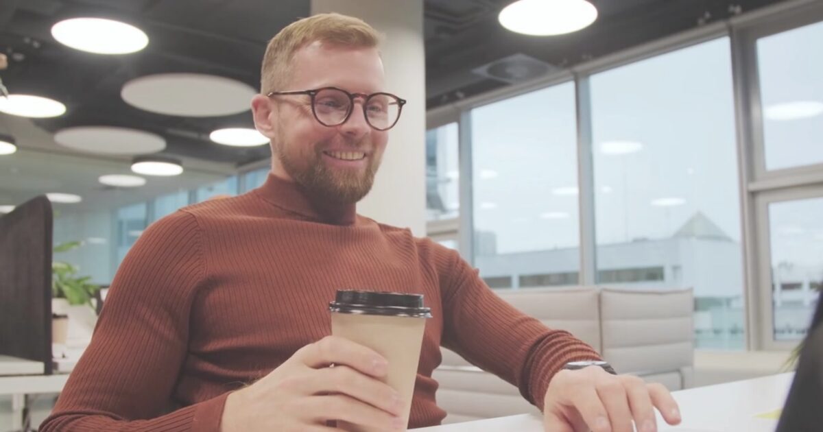 A Caucasian man in a burnt orange sweater, sitting at a desk in a bright office with floor to ceiling windows.