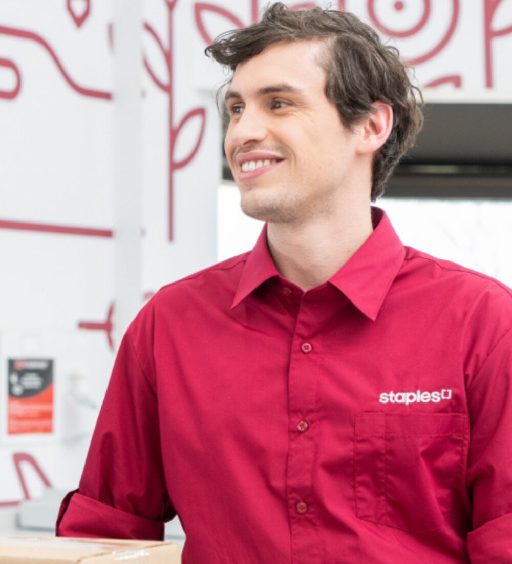 A Young male Caucasian Staples associate smiling in a red Staples shirt.