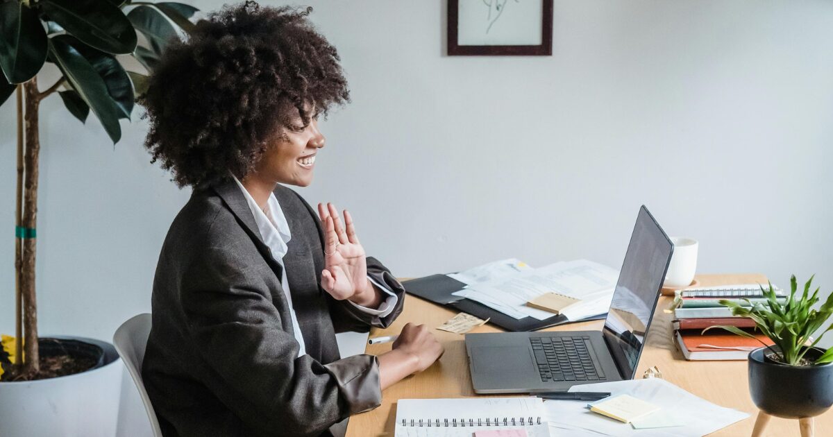 A young Black woman on a video call, sitting at her desk, waving to the camera.