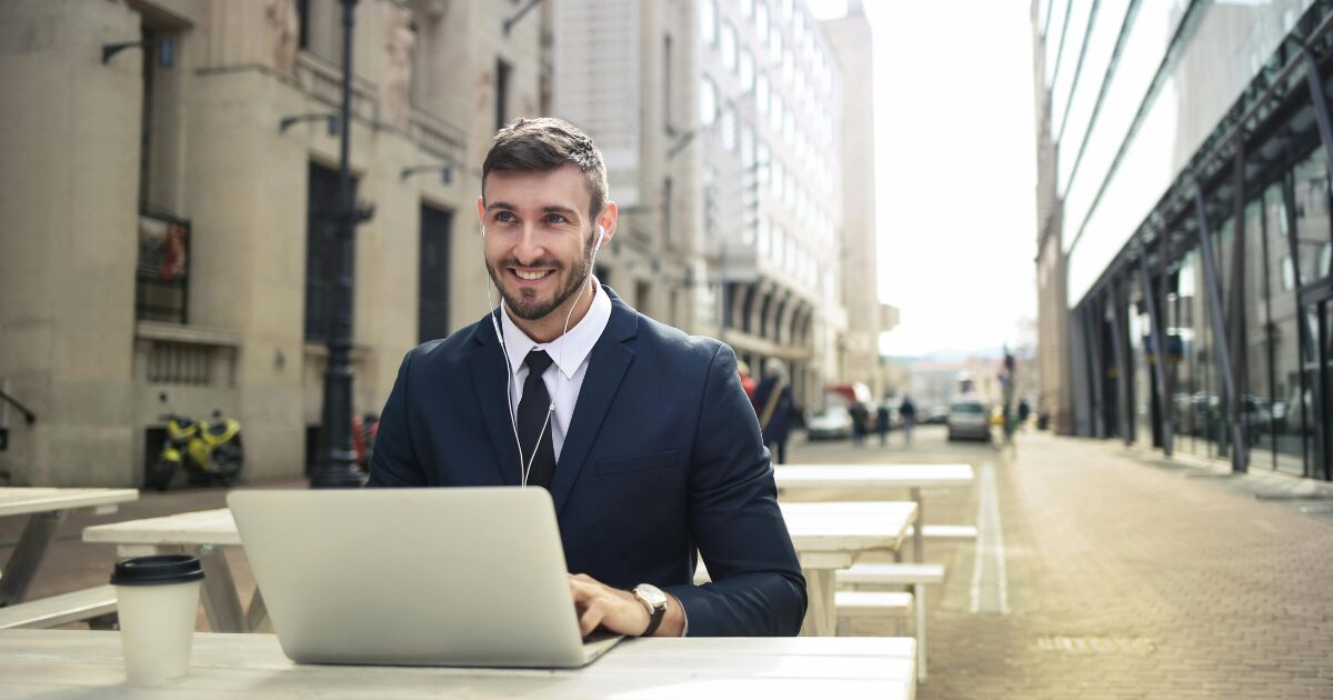 A young Caucasian man in a dark blue suit, sitting outside at a table while typing on his laptop.