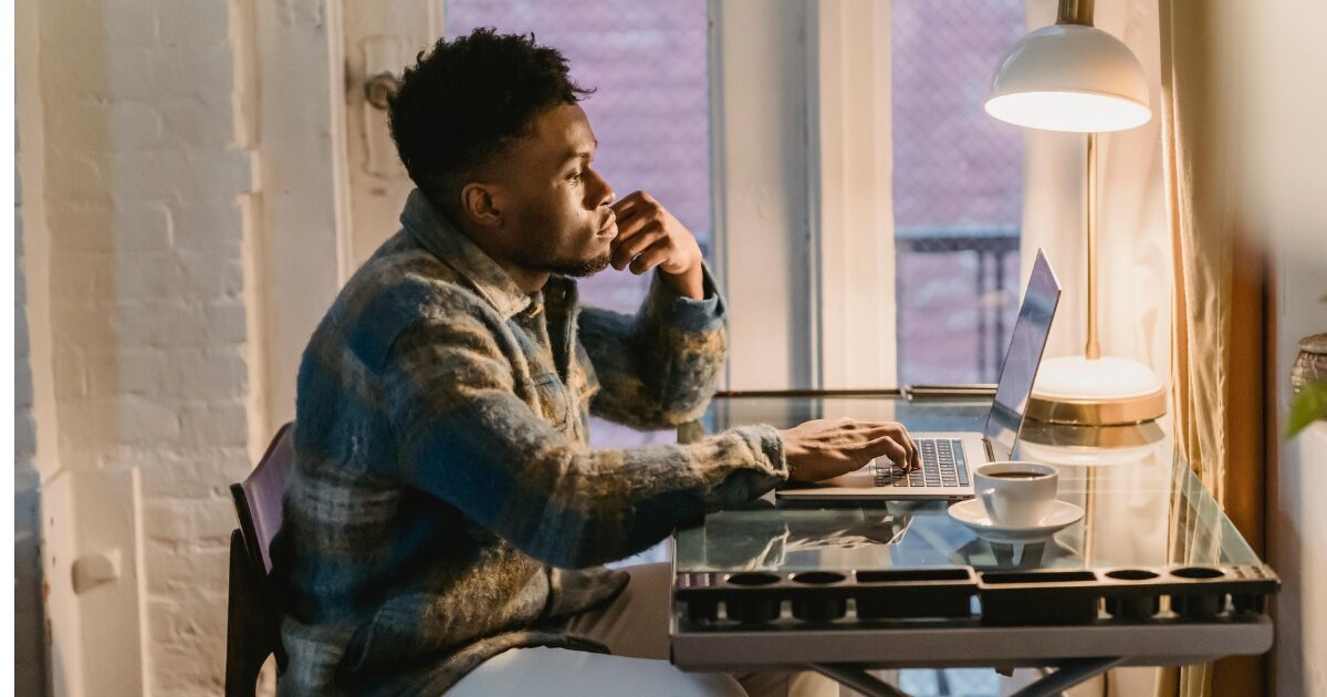 Black man in a blue plaid shirt, sitting at a laptop, at a glass desk.