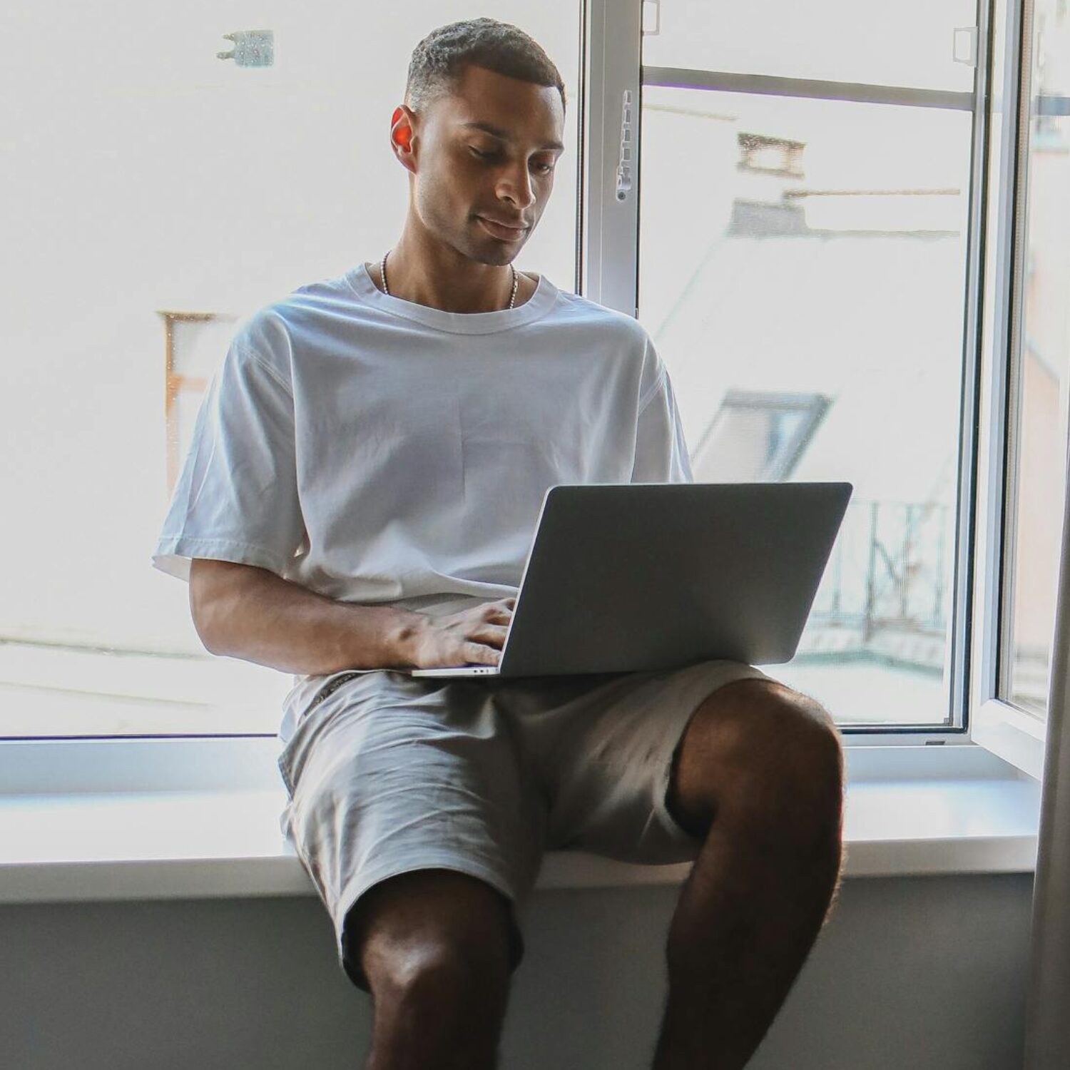 A young Black man sitting on a windowsill on a sunny day, with an open laptop in his lap. He is wearing a white t-shirt.