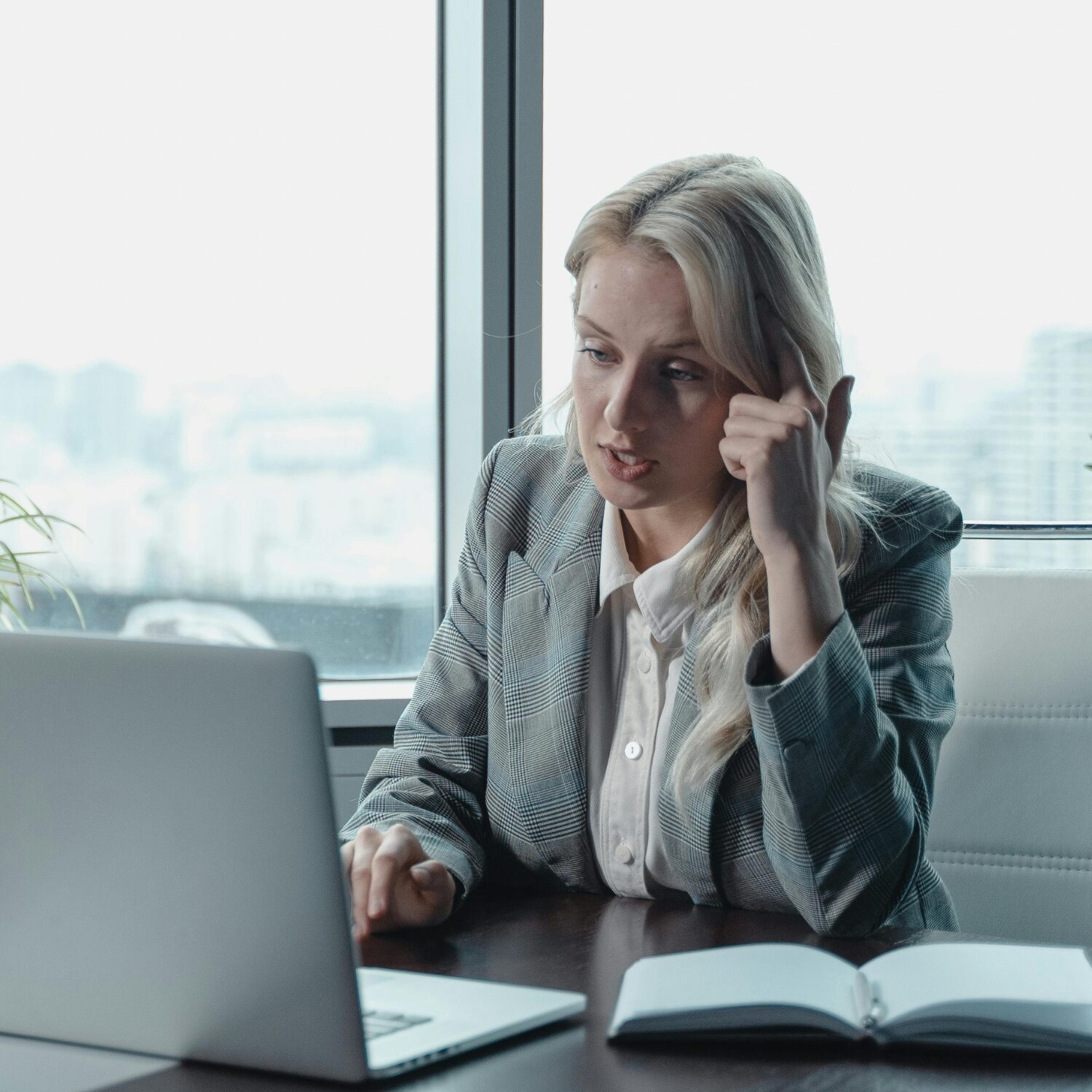Caucasian woman wearing a grey blazer, sitting at a desk, taking a call on her laptop.