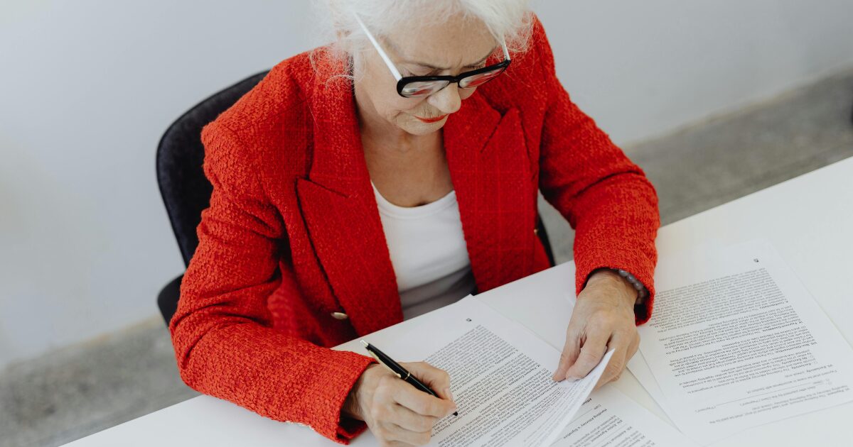 209 An elderly Caucasian woman wearing a red blazer, sitting at a white table, signing a document.