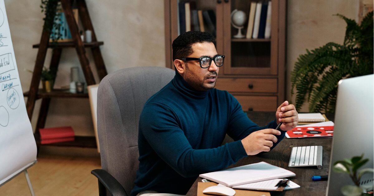 210 A Middle-Eastern man wearing a teal turtleneck, sitting at a computer desk with a pen in his hand and a document in front of him.
