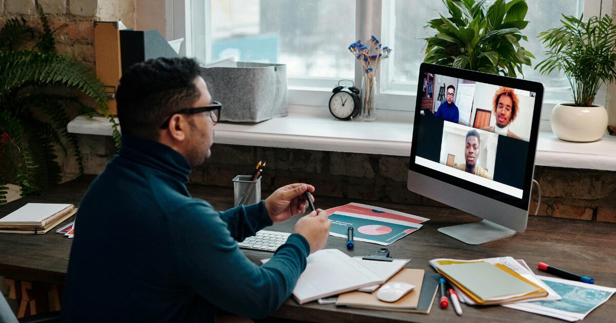 211 A Middle-Eastern man wearing a teal turtleneck, with his laptop open while he's on a video call with 3 other people.
