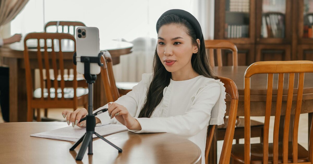 212 A young East Asian woman wearing a white shirt, sitting in front of a tripod that's holding her phone, with a document in front of her and a pen in her hand.