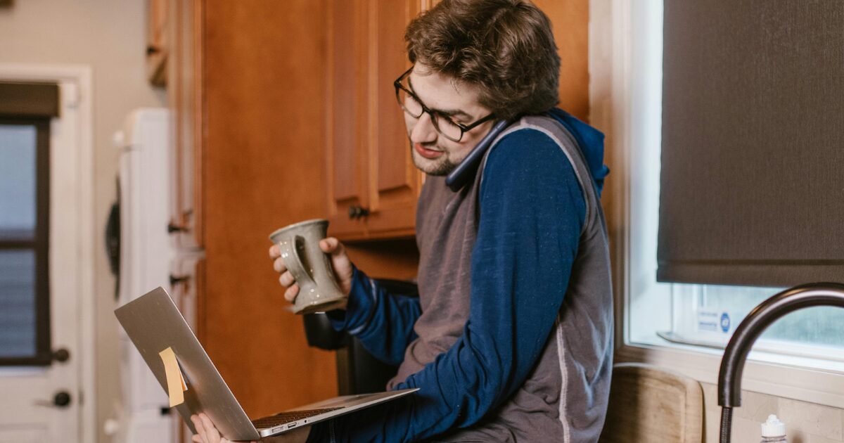 215 A young Caucasian man wearing a hoodie, sitting on a kitchen counter with a coffee cup in his hand and a laptop in his lap.
