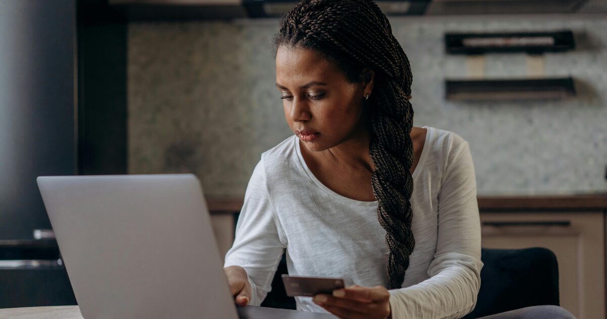 216 A Black woman sitting at a table, entering payment information into her laptop as she holds a card in her other hand.