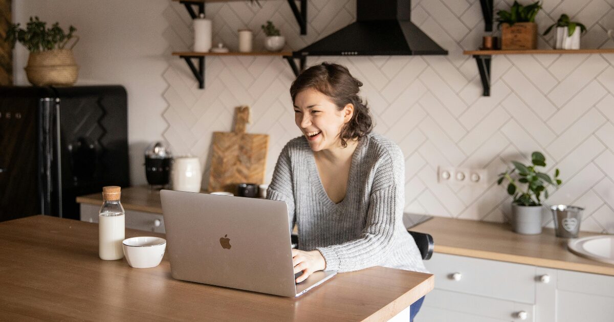 217 Caucasian woman sitting at a wood desk in her kitchen, smiling as she sits in front of her open laptop.