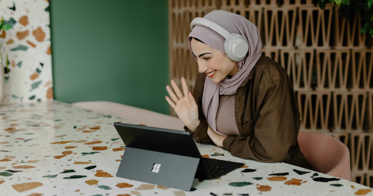 218 A Middle-Eastern woman wearing a grey hijab, sitting at a table as she waves to someone during a video call from her laptop.