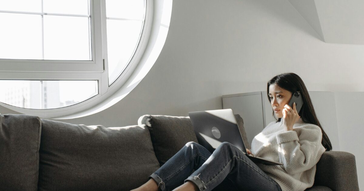 220 East Asian woman wearing a grey sweater, sitting on a couch with an open laptop in her lap.