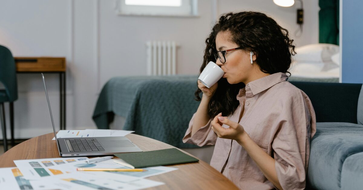 A Black woman sitting at a wood table in front of her laptop, taking a sip of her coffee.