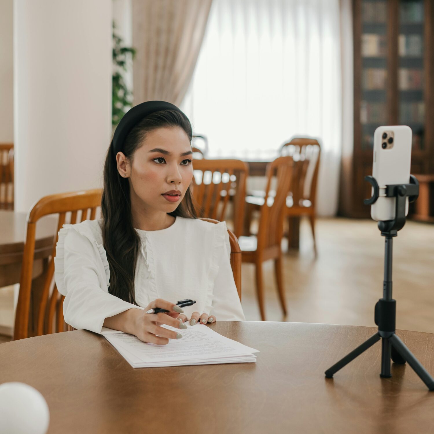 A young East Asian woman wearing a white shirt, sitting in front of a tripod that's holding her phone, with a document in front of her and a pen in her hand.