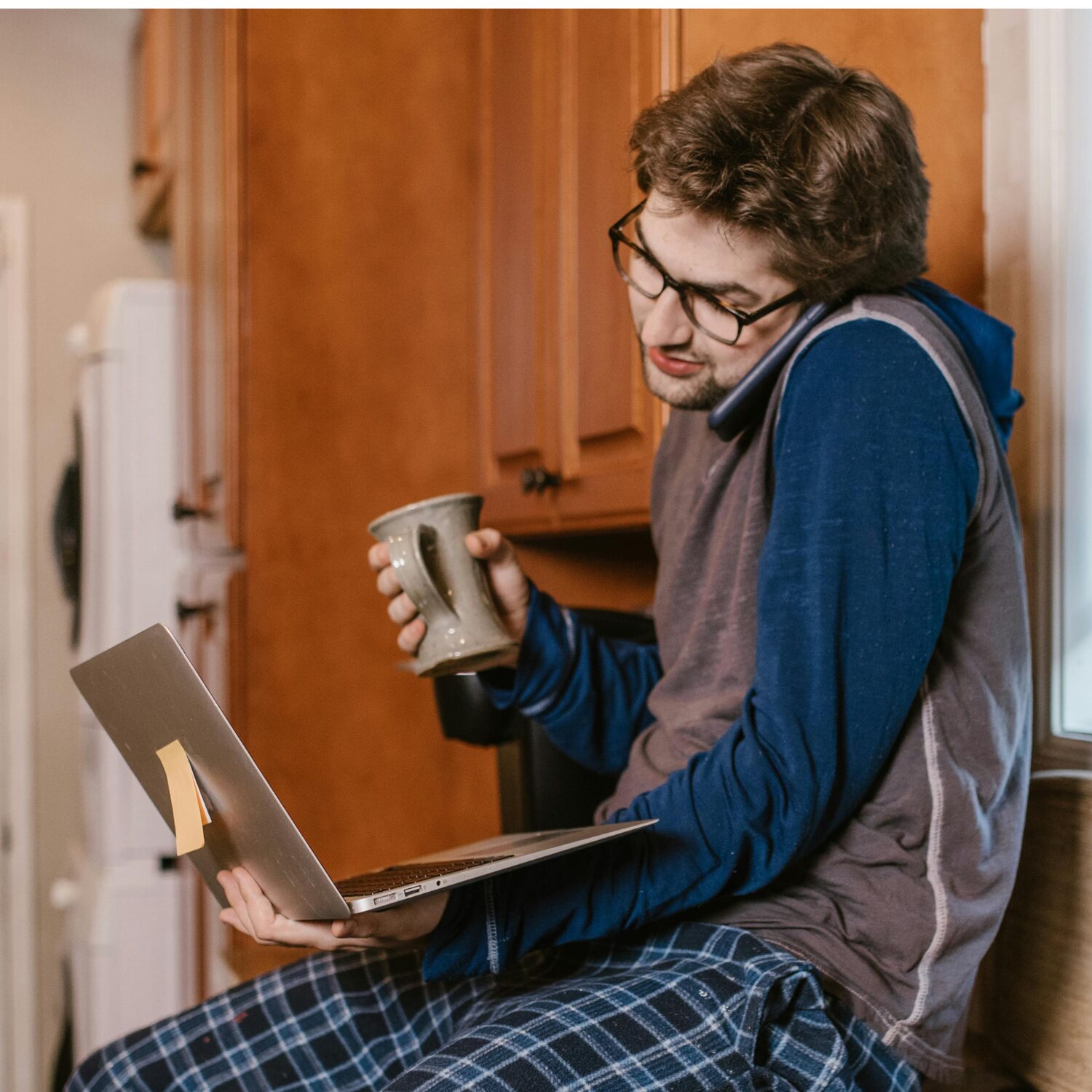 A young Caucasian man wearing a hoodie, sitting on a kitchen counter with a coffee cup in his hand and a laptop in his lap.