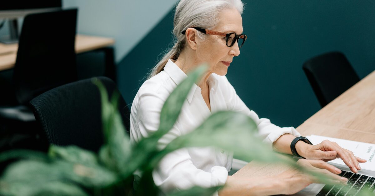 In-Blog Thumbnails (88) Senior Caucasian woman wearing a white button-up shirt, typing on a laptop while sitting at a wood desk.