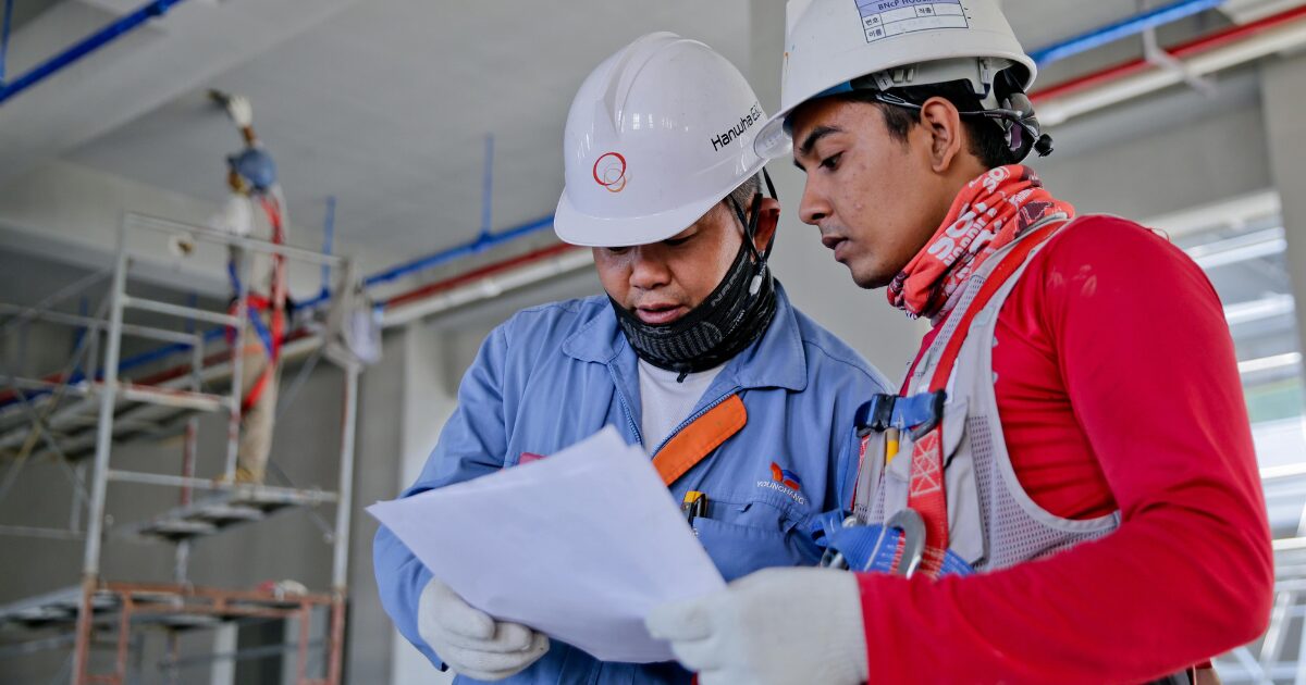 230 Two Latin American male construction workers reading a document. They're both wearing hard hats and construction clothing.