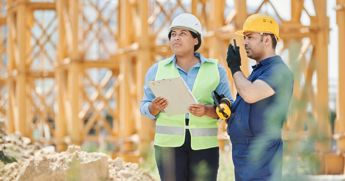 231 An African woman and American man standing on a construction site. The African woman is holding a paper while the American man was talking to a business radio.