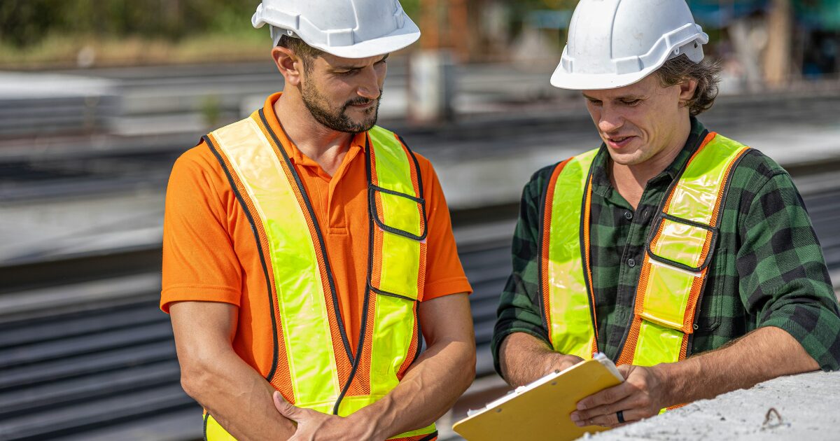 232 Two American male construction workers discussing a document at a construction site. They're wearing hard hats and high-visibility safety vests.